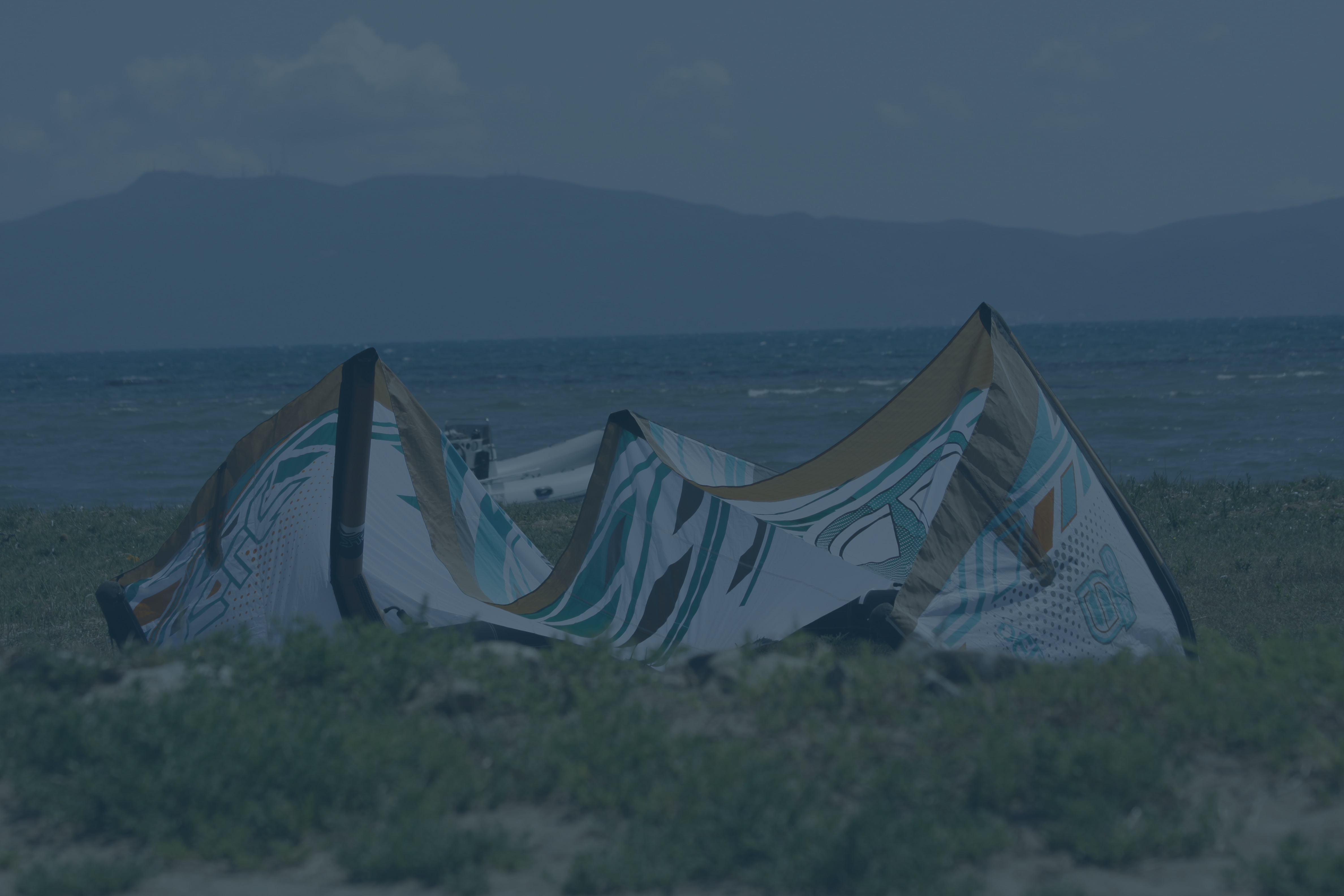 An image of my old kite. There is a grassy beach in the foreground and water with a few white waves in the background. My kite waits upside down on the beach for wind. Behind the kite there is a small boat at the coastline.